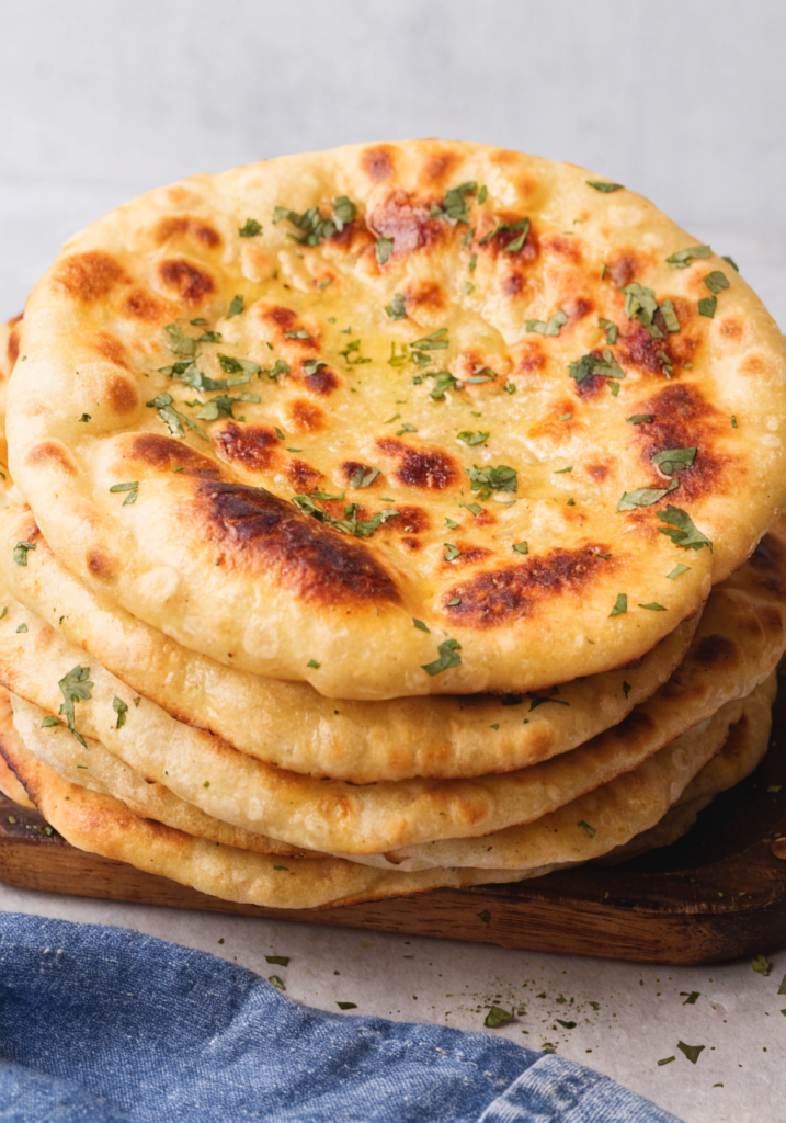 A close-up, slightly high-angle shot features a tall stack of freshly made, golden-brown flatbreads, likely naan or similar leavened bread, resting on a dark wooden board. The top piece of bread is richly textured with golden-yellow color, glistening slightly, and speckled with charred, reddish-brown spots indicating it was cooked at high heat, perhaps in a tandoor or on a griddle. It is generously garnished with finely chopped green herbs, likely cilantro or parsley. Several layers of identical flatbreads are stacked underneath, showing soft, slightly uneven edges. The background is a muted, light gray or off-white texture, providing a soft contrast to the warm tones of the bread. A sliver of a blue, textured cloth (possibly denim or linen) is visible in the lower foreground.